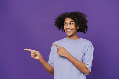 Young happy smiling handsome latin curly man with piercing pointing and looking aside while standing over isolated violet background