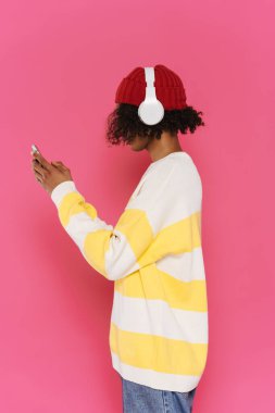Young caribbean man in headphones using mobile phone isolated over pink background