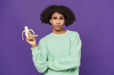 Young caribbean man frowning while eating banana isolated over purple background