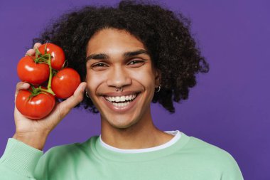 Young caribbean man smiling while showing tomatoes isolated over purple background
