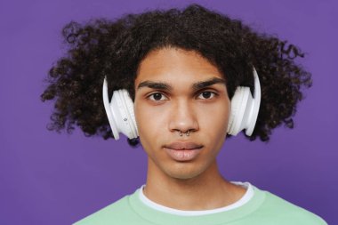 Young caribbean man with piercing listening music with headphones isolated over purple background