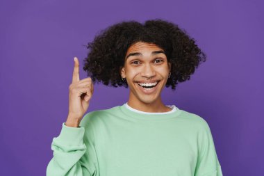 Young caribbean man smiling and pointing finger upward isolated over purple background