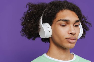 Young caribbean man with piercing listening music with headphones isolated over purple background