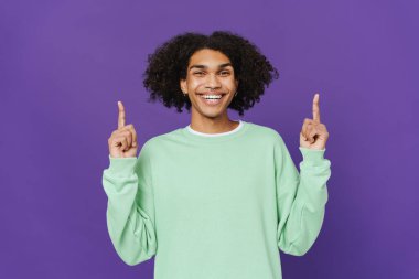 Young caribbean man smiling and pointing fingers upward isolated over purple background
