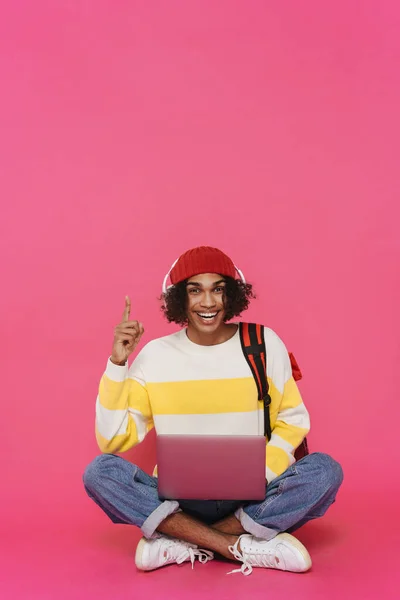 Black man in headphones pointing finger upward and using laptop isolated over pink background