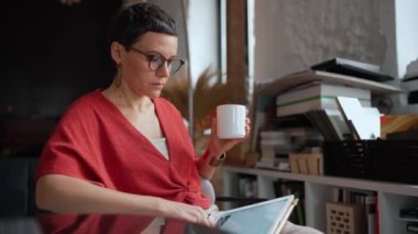 Serious woman architect with short hair in eyeglasses working on tablet and drinking coffee in the office