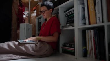 Serious brunette woman architect in eyeglasses working on tablet while sitting on the floor in the office