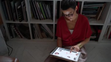 Confident brunette woman architect in eyeglasses working on tablet while sitting on the floor in the office