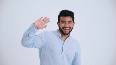 Smiling Indian man with piercing wearing blue shirt waving hand at the camera in the grey studio
