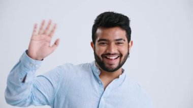Laughing Indian man with piercing wearing blue shirt waving hand at the camera in the grey studio