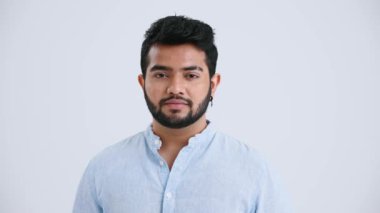 Serious Indian man with piercing wearing blue shirt opening eyes at the camera in the grey studio