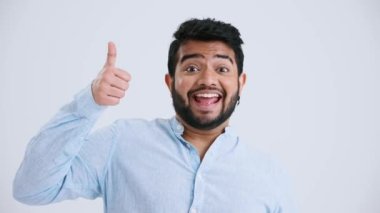 Smiling Indian man with piercing wearing blue shirt showing thumbs up at the camera in the grey studio