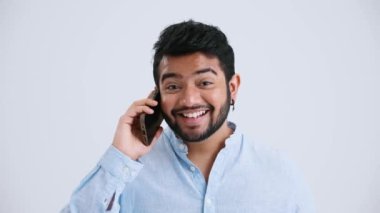 Rejoicing Indian man with piercing wearing blue shirt talking on phone in the grey studio