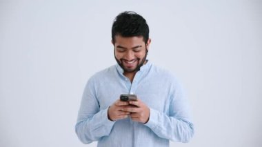 Laughing Indian man with piercing wearing blue shirt texting on phone in the grey studio