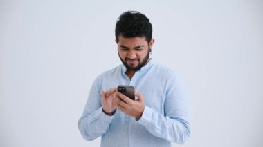 Handsome Indian man with piercing wearing blue shirt texting on smartphone in the grey studio