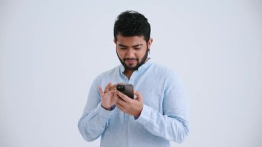 Cute Indian man with piercing wearing blue shirt typing on smartphone in the grey studio