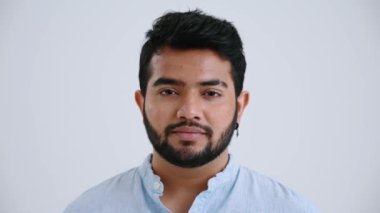 Concentrated Indian man with piercing wearing blue shirt opening eyes at the camera in the grey studio