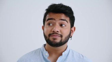 Shocked Indian man with piercing wearing blue shirt looking around and showing silence gesture at the camera in the grey studio