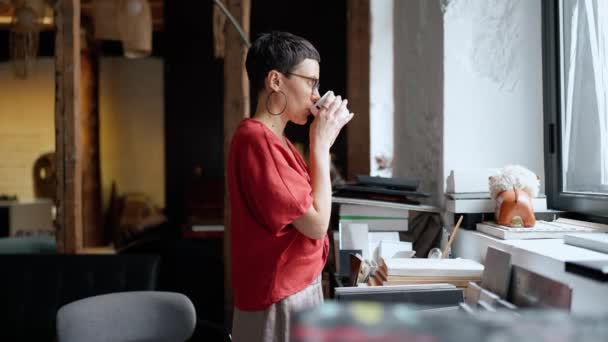 自信を持ってブルネットの女性建築家は窓を見てオフィスでコーヒーを飲む — ストック動画