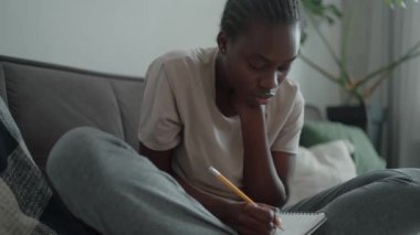 Concentrated African woman with pigtails and depression writing something in notepad on the couch at home