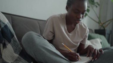 Confident African woman with pigtails and depression writing something in notepad on the couch at home