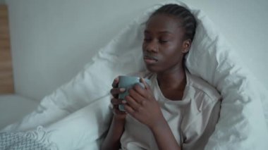 Bored African woman with pigtails and depression drinking cup of tea on the bed at home