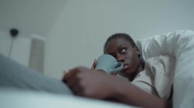 Bored African woman with pigtails and depression reading book and drinking cup of tea on the bed at home
