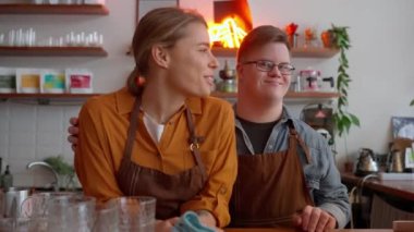 Smiling man barista with Down syndrome listening and hugging his girl colleague in a cafe