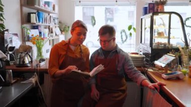 Concentrated man barista with Down syndrome reading book and talking with his girl colleague in a cafe