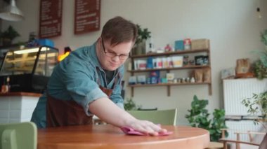 Positive man barista with Down syndrome wiping table in cafe