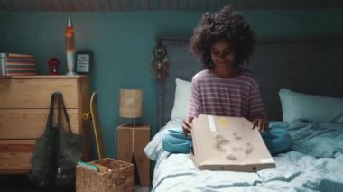 Smiling curly haired young African girl opening pizza in the box on the bed at home