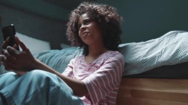 Happy curly haired young African girl talking by video call on phone near bed at home