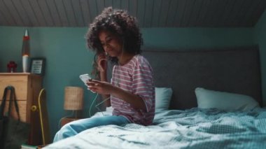 Side view of curly haired young African girl typing on phone in headphones on the bed at home