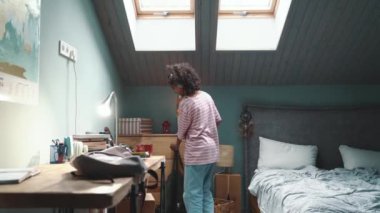 Smiling curly young African girl listening to music and packing her backpack at home