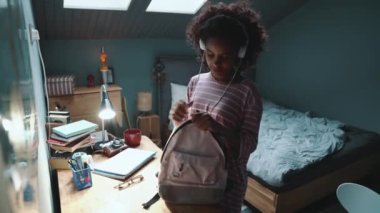 Positive curly young African girl listening to music and closes the zipper on the backpack at home