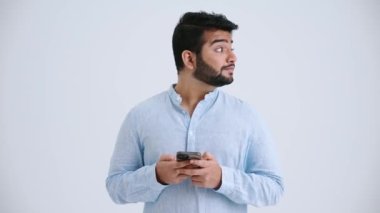 Happy Indian man with piercing wearing blue shirt typing on mobile and looking around in the grey studio