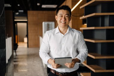 Portrait of smiling cheerful positive asian businessman in white shirt with tablet looking directly in camera indoor