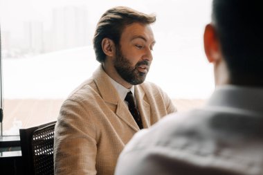 Adult white businessman talking to his male partner while sitting by table indoors