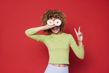 Young curly woman with opened mouth holding donuts on her eyes and showing victory gesture , while standing over red isolated background