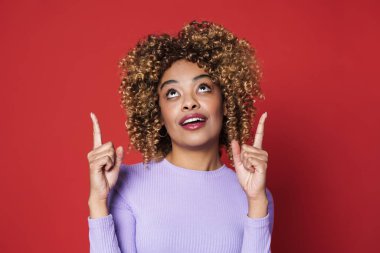 Young beautiful attractive curly woman pointing and looking up over isolated red background
