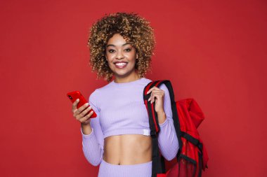 Young black woman with backpack smiling and holding cellphone isolated over red background