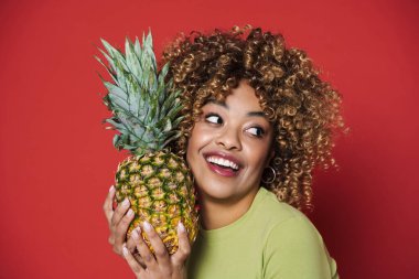 Young black woman laughing while posing with pineapple isolated over red background
