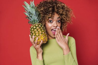 Young black woman expressing surprise while posing with pineapple isolated over red background