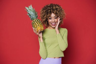 Young black woman laughing while posing with pineapple isolated over red background