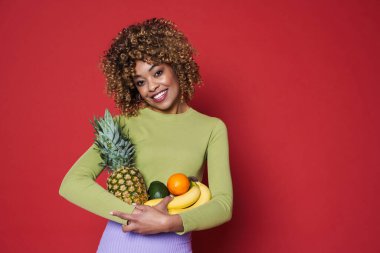 Young black woman laughing while posing with fruits isolated over red background