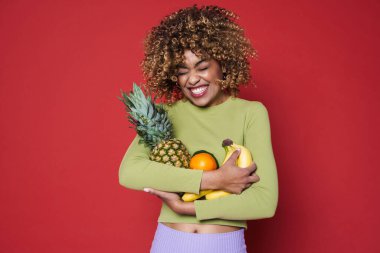 Young black woman laughing while posing with fruits isolated over red background