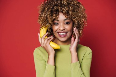 Young black woman laughing while posing with banana isolated over red background