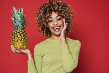 Young black woman laughing while posing with pineapple isolated over red background