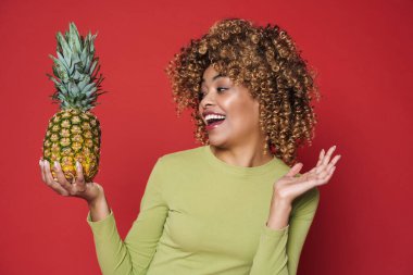 Young black woman laughing while posing with pineapple isolated over red background
