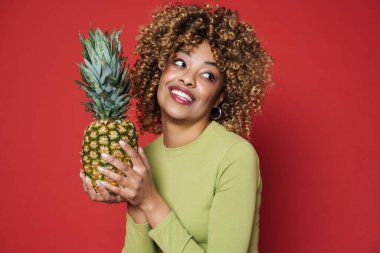 Young black woman laughing while posing with pineapple isolated over red background
